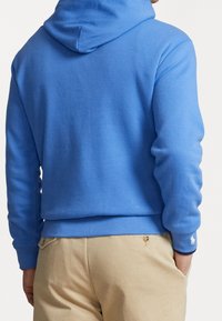 Man med ljusblå hooded sweatshirt och beige byxor, händerna i fickorna, vy från ryggen och midjan upp.