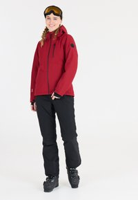 Röd isolerad jacka med huva och dragkedja, kombinerad med svarta snöbyxor och svarta skidor. Funktioner inkluderar en logotyp patch och goggles.