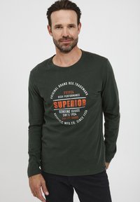 Mørkegrønt langærmet T-shirt med et vintage print og orange og hvid tekst. Lavet af bomuld, har det rund hals og en let figursyet pasform.