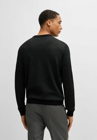 Maglione nero a maglia con texture a coste e scollo rotondo, con polsini e orlo neri a contrasto. Modello che indossa pantaloni grigi.
