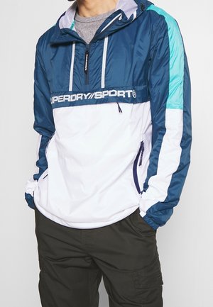 Mand iført en blå, hvid og teal Superdry Sport vindjakke med frontlynlås og sidelommer, kombineret med mørkegrønne cargobukser.