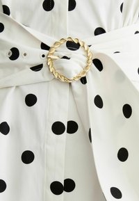 Tejido blanco con grandes lunares negros recogido y asegurado por una hebilla circular torcida de color dorado.