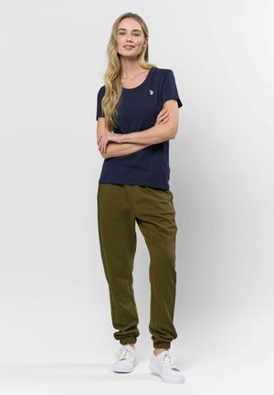 Jūras zila t-krekls no mīksta auduma ar īsām piedurknēm un apaļu kakla izgriezumu, kombinēts ar olīvu zaļiem sporta biksēm ar elastīgiem aprocēm un baltām kedām.