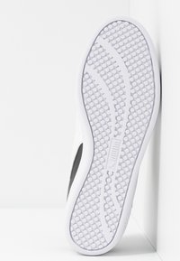 Semelle d'une basket blanche avec un motif d'adhérence circulaire et deux lignes de traction courbées, positionnée verticalement contre un mur d'angle blanc.