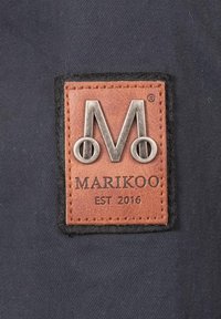 Ruda odinė etiketė stačiakampio formos su sidabriniu metaliniu logotipu "M" ir tekstu "MARIKOO EST 2016", siuvinėta tamsiame audinio fone.