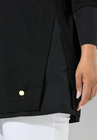 Schwarze Langarm-Tunika mit seitlichen Schlitzen, weichem Stoff und einem kleinen goldenen Logo-Akzent am Saum, kombiniert mit weißen Hosen.