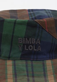 Sombrero tipo bucket hecho de tela estampada en verde, marrón y azul. Presenta un logotipo bordado "BIMBA Y LOLA" en la banda.