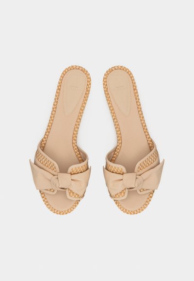 Μπεζ σανδάλια τύπου slip-on με πλεκτή διακόσμηση και μεγάλα υφασμάτινα φιόγκους στο μπροστινό μέρος, τοποθετημένα δίπλα δίπλα σε λευκό φόντο.