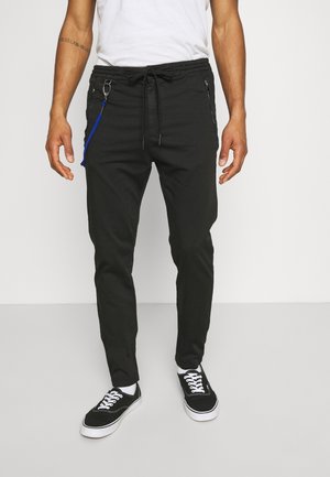 Mand iført sorte slim fit bukser med bindebånd i taljen, blå nøglering, hvide sokker, sorte og hvide sneakers og en hvid t-shirt.