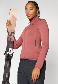 En kvinne med flettet hår har på seg en rosa zip-up jakke og holder et ski med bindinger mot en nøytral bakgrunn.
