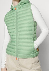 Jasnozielona kamizelka puchowa z kapturem, z poziomym pikowaniem, zamek z przodu i pomarańczowy uchwyt suwaka.
