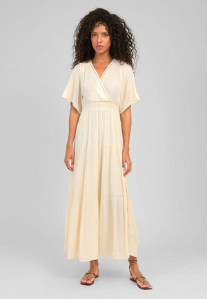 Femme aux cheveux bouclés portant une robe crème à manches courtes et longueur cheville, ainsi que des sandales plates marron à brides, debout devant un fond gris.