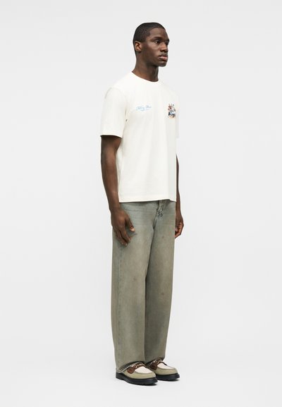 Μαύρος άνδρας που στέκεται, φοράει λευκό μπλουζάκι με μικρό γραφικό, φαρδιά γκρι τζιν και μπεζ και καφέ μοκασίνια, μπροστά σε απλό λευκό φόντο.