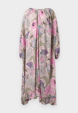 Femme portant une longue robe fleurie ample dans des tons rose, violet et beige, debout, une main sur la hanche, et des sandales noires à talons hauts.