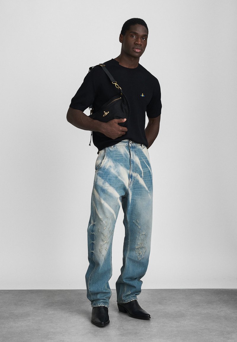 Svart kortärmad skjorta med en logotyp, slitna ljusblå jeans med tie-dye-mönster, håller en svart väska och har på sig svarta spetsiga ankelstövlar.