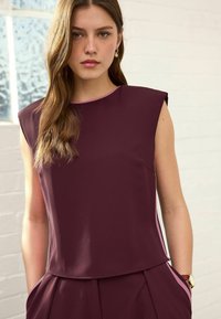 Burgundy sleeveless top with a round neckline and subtle pink trim. Smooth fabric, relaxed fit, paired with matching high-waisted trousers.
