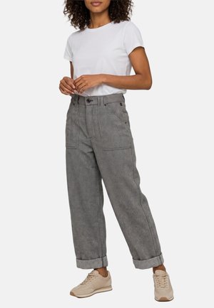 Donna con camicia bianca a maniche corte, pantaloni grigi a vita alta, a gamba larga con risvolti e sneakers beige, in piedi con le mani incrociate.