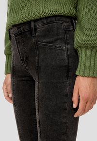 Czarne dżinsy o slim fit, z pięcioma kieszeniami, subtelnym przeszyciem i srebrnym guzikiem, w połączeniu z zielonym, teksturowanym swetrem.