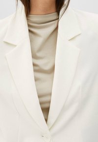 Biały żakiet z klapami notch i pojedynczym zapięciem na guzik, nałożony na beżowy top z wysokim kołnierzem. Teksturowana tkanina, dopasowany krój.