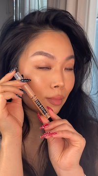 NYX Pro Finish Stick, przezroczysta tubka, jasnobeżowy produkt, gładka tekstura. Modelka ma długie ciemne włosy, trzyma produkt, paznokcie w czerwone i białe kropki.