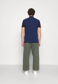 Marinblå polo med korta svarta ärmar, med en grön detalj på kragen och en logotyp patch på vänster ärm. Olivgröna vida byxor.