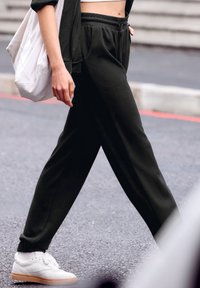 Schwarze Jogginghose aus weichem Material, mit elastischem Bund und seitlichen Taschen, kombiniert mit weißen Sneakers und einer leichten Tasche.