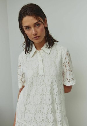 Vestido tipo polo de encaje blanco con patrones florales, mangas cortas y un cuello abotonado con acabado texturizado.