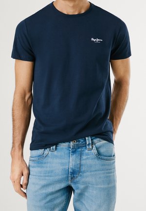 T-shirt en coton navy avec col rond et manches courtes. Présente un petit logo blanc sur la poitrine, associé à un jean en denim bleu clair.