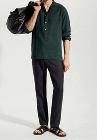 Camicia di lino verde scuro con colletto a camicia, abbinata a pantaloni scuri e sandali neri. Una borsa nera è appoggiata su una spalla.