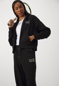 Črna zip-up kapuca z sprednjimi žepi, v kombinaciji z usklajenimi trenirkami, ki imajo zlate vezenine logotipa. Model drži teniško lopar.
