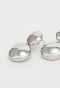 Sølvfargede øredobber med en glatt, oval form. Har en polert overflate med et preget "BOSS"-logo. Enkel, moderne design.