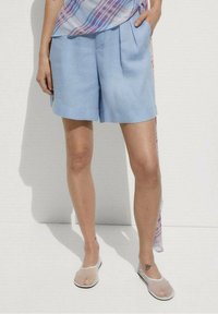 Pantalones cortos de lino azul claro con un ajuste holgado, parte delantera lisa y pliegues sutiles. Combinados con una blusa estampada multicolor y sandalias transparentes.