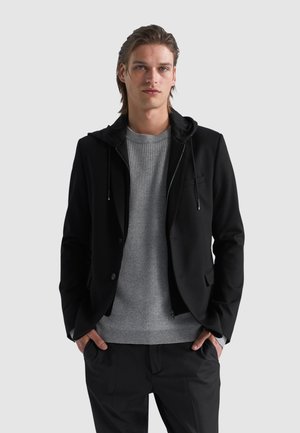 Giacca nera con dettaglio della felpa con cappuccio, abbinata a un maglione grigio testurizzato. Il modello sta in piedi con le mani in tasca, indossando pantaloni neri.