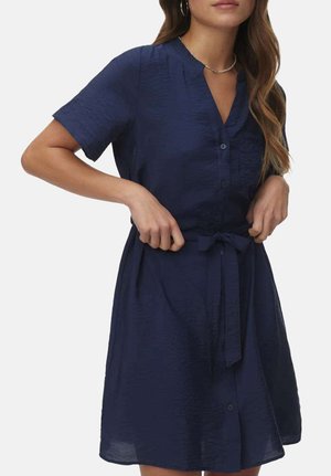 Femme portant une robe bleu marine à manches courtes avec une patte de boutonnage à l'avant, nouée à la taille par une ceinture en tissu, visible des épaules jusqu'au milieu des cuisses.