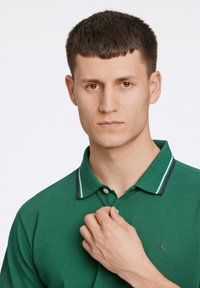 Zielona koszulka polo z kołnierzykiem, ozdobiona białymi i fioletowymi paskami. Teksturowana tkanina oraz wyszywane logo po lewej stronie.