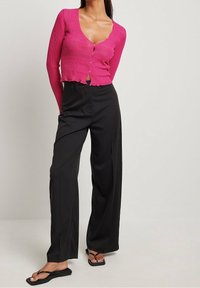 Eng anliegender, gerippter rosa Cardigan mit tiefem Ausschnitt und Knopfleiste, kombiniert mit hoch taillierter, schwarzer, weit geschnittener Hose und flachen Sandalen.