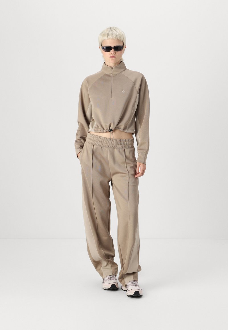 Maglione beige con zip e colletto alto, abbinato a pantaloni beige larghi. Entrambi hanno una texture liscia e dettagli di design sottili.