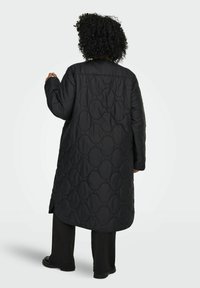 Juoda ilga siūta striukė su bangelių raštu, laisvo stiliaus ir šoninėmis skeltukėmis. Medžiaga atrodo lygi ir lengva.