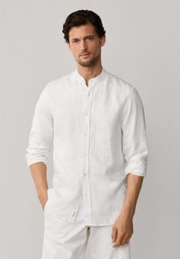 Hombre de cabello oscuro con camisa blanca de manga larga y cuello mandarín y pantalones blancos, posando con una mano en el bolsillo sobre un fondo liso.