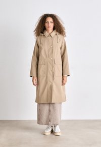 Beige regenjas met een capuchon, gouden hardware, twee voorzakken en een trekkoorddetail. Gecombineerd met een lange beige rok en witte sneakers.