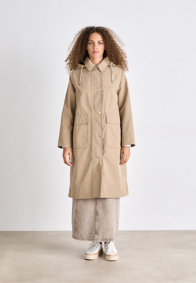 Beige regenjas met een capuchon, gouden hardware, twee voorzakken en een trekkoorddetail. Gecombineerd met een lange beige rok en witte sneakers.