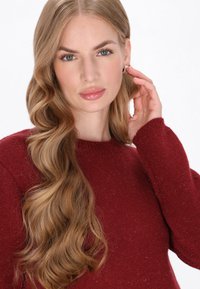 Jersey de punto rojo con cuello redondo y acabado texturizado. Cabello largo y ondulado, maquillaje sutil y pendientes visibles.