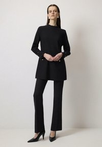 Túnica negra de mangas largas, con detalles dorados en los puños, combinada con pantalones negros ajustados y tacones negros afilados. Textura suave.