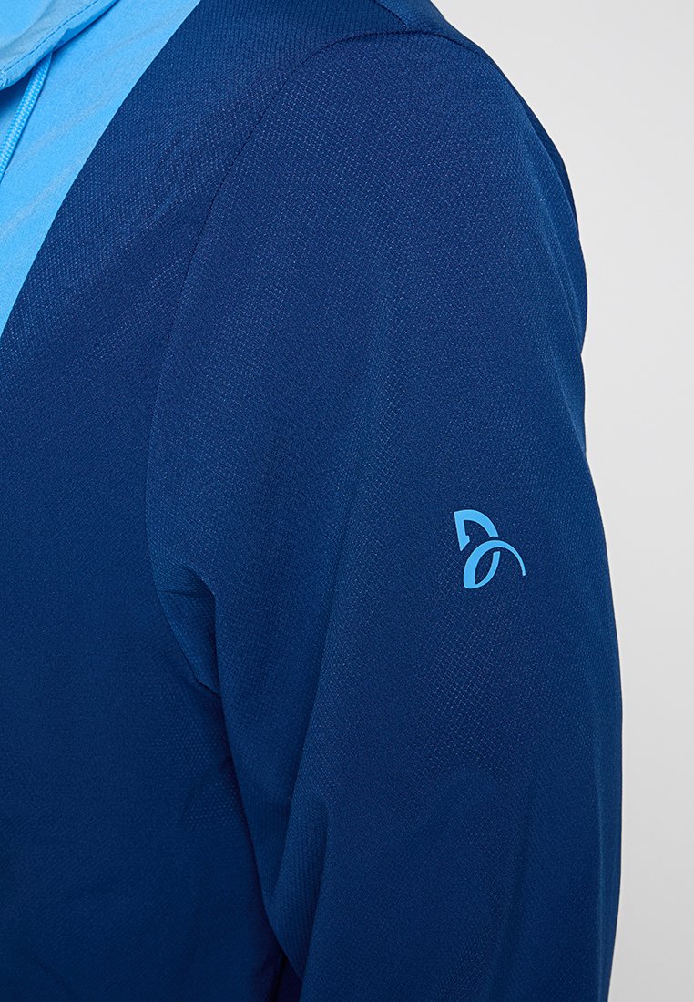 Primo piano di una persona in giacca blu navy con texture e un logo astratto blu chiaro sulla manica superiore.