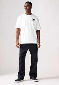 T-shirt blanc à manches courtes avec un logo cartoon noir sur la poitrine, associé à un pantalon de survêtement noir et des chaussures foncées. Tissu lisse et décontracté.