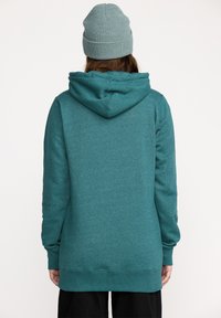 Personne portant un hoodie teal et un bonnet en laine bleu clair, se tenant de dos contre un fond blanc uni.