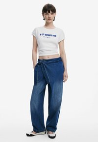 Vit cropped t-shirt med blå text, kombinerad med högmidjade blå denimbyxor med knytbälte. Enkla svartvita skor.