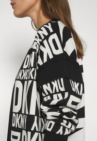 Svart och vit stickad tröja med stor, djärv textgrafik. Texturerna varierar mellan släta och ribbade. Oversized passform med sänkta axlar.