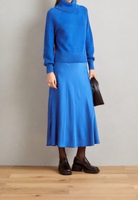 Sininen neulepaita, jossa on teksturoidut hihat, yhdistetty virtaavaan siniseen satiinishortsiin ja mustiin paksupohjaisiin kenkiin. Korostettu mustalla clutch-laukulla.