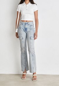 Kvinna som bär ljusblå cargo-jeans med slitsar, vit kortärmad polo-tröja, vita sandaler och bär en vit axelväska.